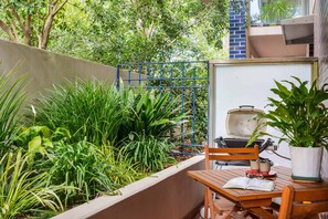 A patio with a wooden table and chairs, a grill, and lush greenery. - Stylish River Boulevard Apartment in Richmond (Richmond)