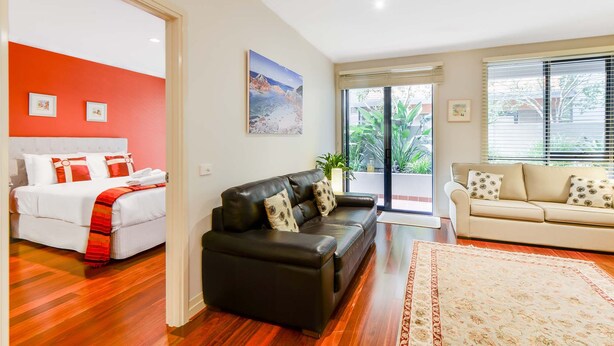 A hotel room with a bed, a black leather sofa, a beige sofa, and a large window with blinds. - Stylish River Boulevard Apartment in Richmond (Richmond)