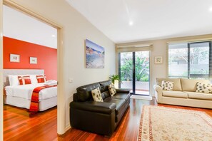 Habitación de hotel con cama, sofá de cuero negro, sofá beige y un ventanal con persianas.
