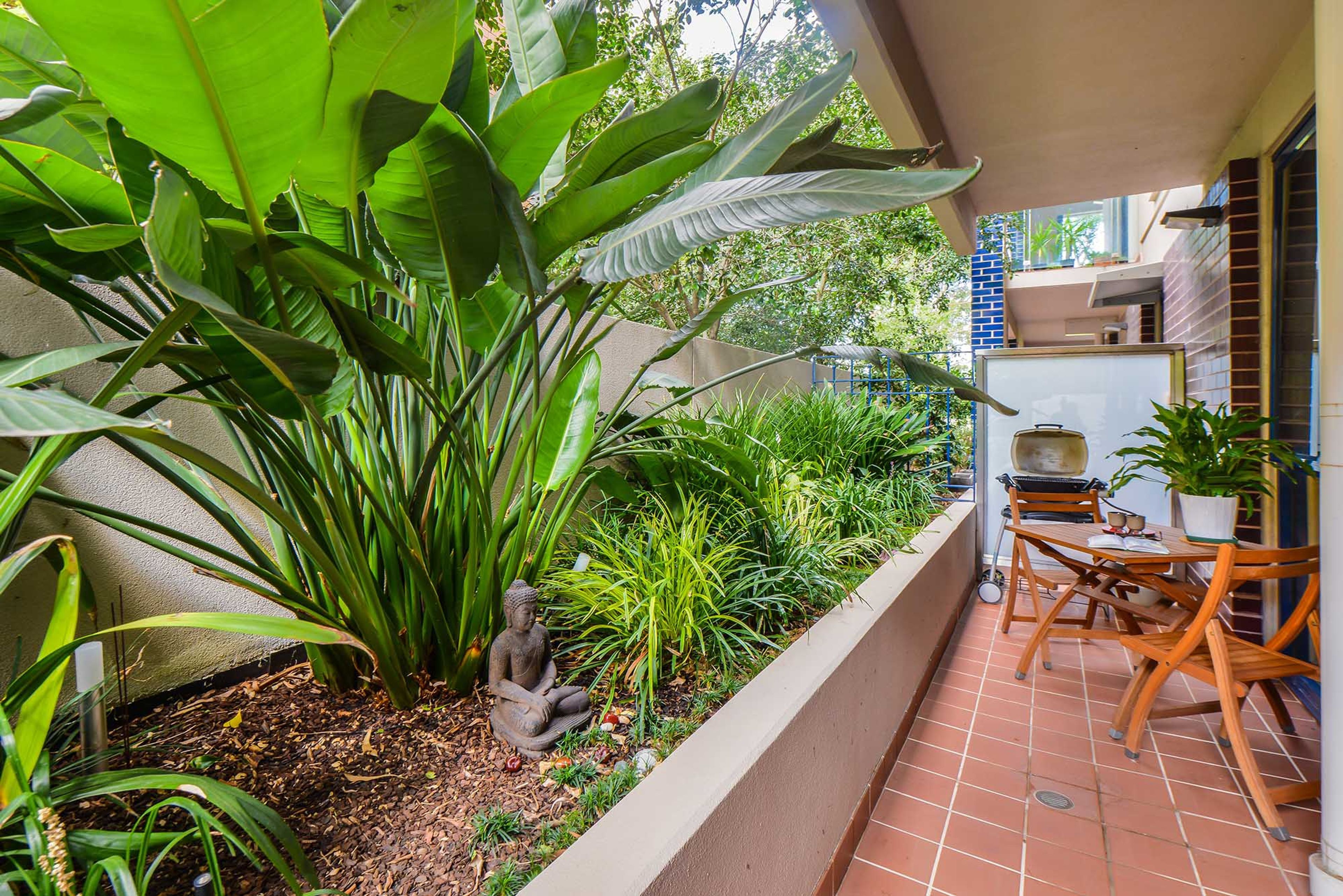 A patio area with a table and chairs, surrounded by various plants and a Buddha statue.