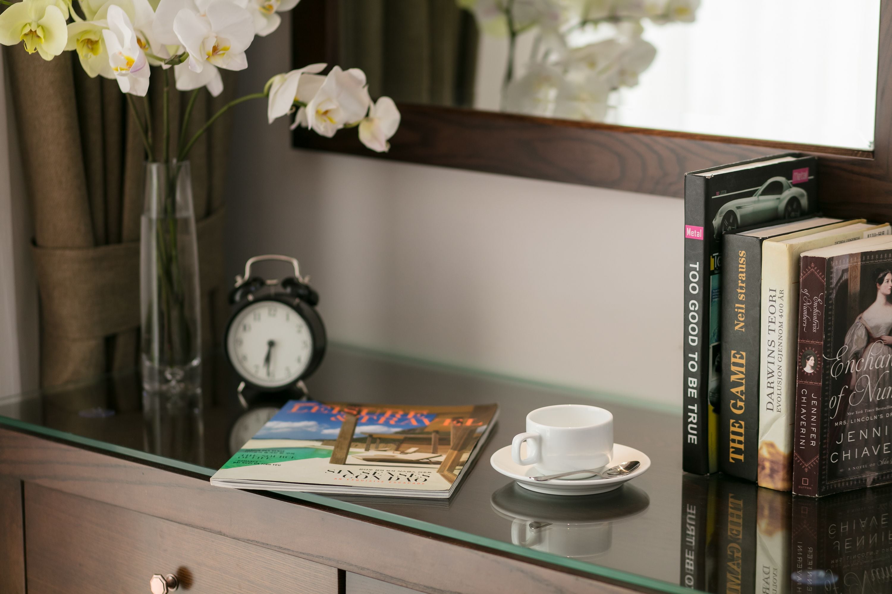 Une table à plateau de verre sur laquelle se trouvent une tasse blanche, un livre intitulé « Le Jeu », une horloge et un vase avec des fleurs blanches.