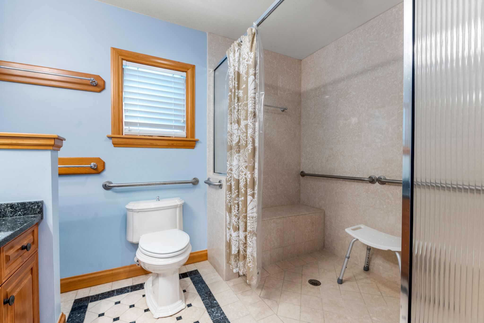 Une salle de bain comprenant des toilettes, une douche avec une porte en verre, une fenêtre avec des stores et une barre d’appui.