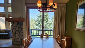 A dining area with a wooden table and chairs, a stone fireplace, and a view of trees through a large window. - Snowcreek Resort Vacation Rental 858 (Mammoth Lakes)