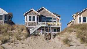 Ein zweistöckiges Strandhaus mit Balkon und einer Treppe zum Eingang, im Hintergrund ein Sandstrand.