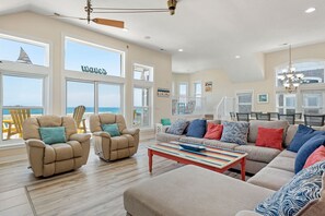 A spacious living room with a large window overlooking the ocean, a cozy seating area with multiple chairs and a sofa, and a staircase leading to an upper level. - Giggling Gecko: Oceanfront, Private Pool, Hot tub (Nags Head)