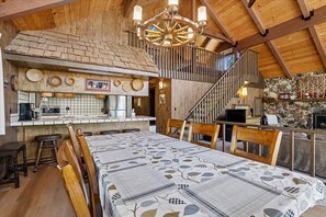 A dining area with a long table, wooden chairs, a chandelier, and a kitchen area with a stone wall and a fireplace.