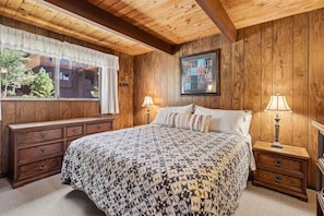 Une chambre avec un plafond en bois, un grand lit avec une literie à motifs, deux tables de chevet avec des lampes, une commode et une fenêtre avec des rideaux.