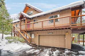 Een houten chalet met twee verdiepingen, een overdekte garage en een houten trap naar de bovenverdieping.
