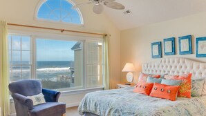 Une chambre avec une grande fenêtre donnant sur l’océan, une tête de lit capitonnée et un lit agrémenté d’oreillers colorés.