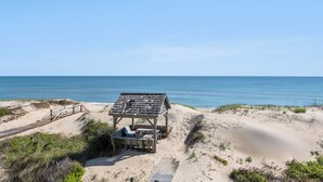 Un abri de plage avec un banc et une table, entouré de dunes de sable et d’une promenade en bois.