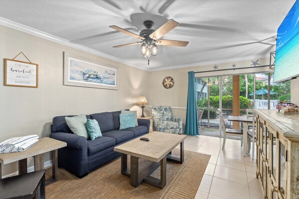 A living room with a sofa, coffee table, and a ceiling fan. There is a window with curtains and a view of greenery outside.