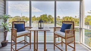 A wooden porch with two chairs and a table, overlooking a tennis court and greenery. - Cedars East #528 Townhome by RVA (Longboat Key)