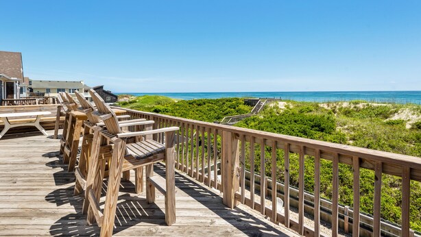 Een houten terras met stoelen en tafels, uitkijkend op een strand en zee.