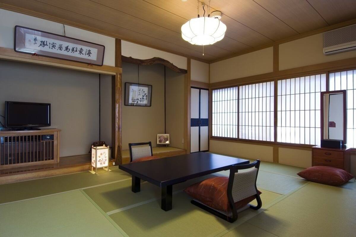 A traditional Japanese room with tatami flooring, a low table, chairs, a television, and a large window with shoji screens.