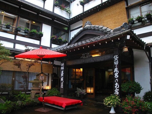 Un bâtiment traditionnel japonais, avec un parasol rouge, une lanterne en pierre et un petit jardin agrémenté d’un banc.