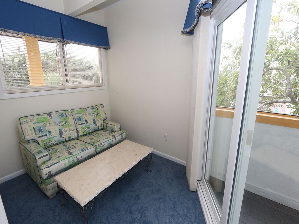 A small room with a blue carpet, a green patterned sofa, a white bench, and a sliding glass door leading to an outdoor area.