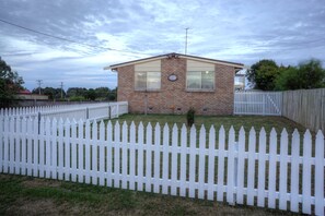 Una casa a un piano in mattoni con una recinzione in legno bianco, un piccolo giardino e un cielo sereno.