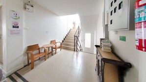 Un espace d’accueil avec un escalier, des chaises en bois, une petite table et un extincteur fixé au mur.