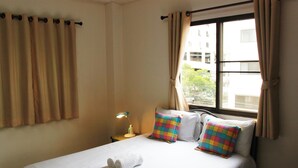 Une chambre d’hôtel avec un lit, deux oreillers colorés, une table de chevet avec une lampe et une fenêtre avec des rideaux.