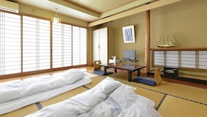 Una habitación japonesa tradicional con suelo de tatami, una mesa baja con una silla, una cama con ropa blanca y un modelo de barco en la pared.