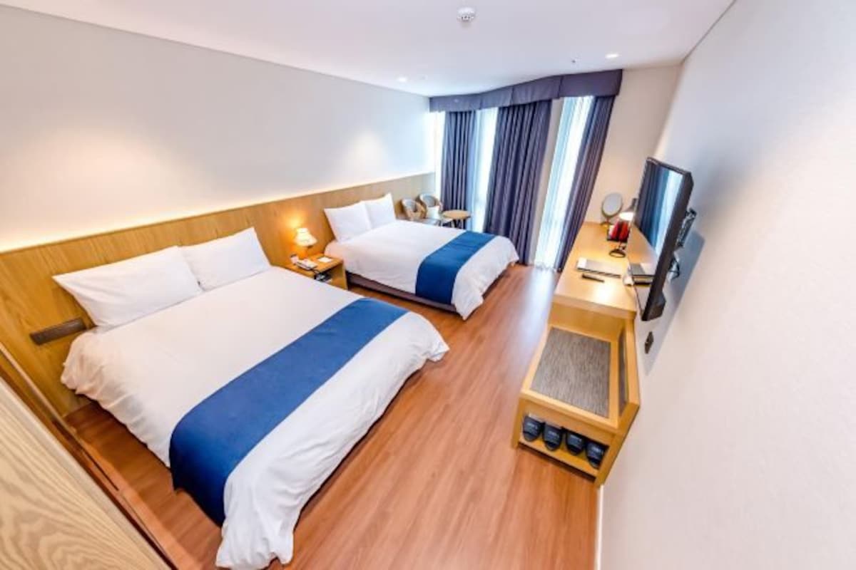 A hotel room with two beds, a TV, a desk with a lamp, and a shoe rack.