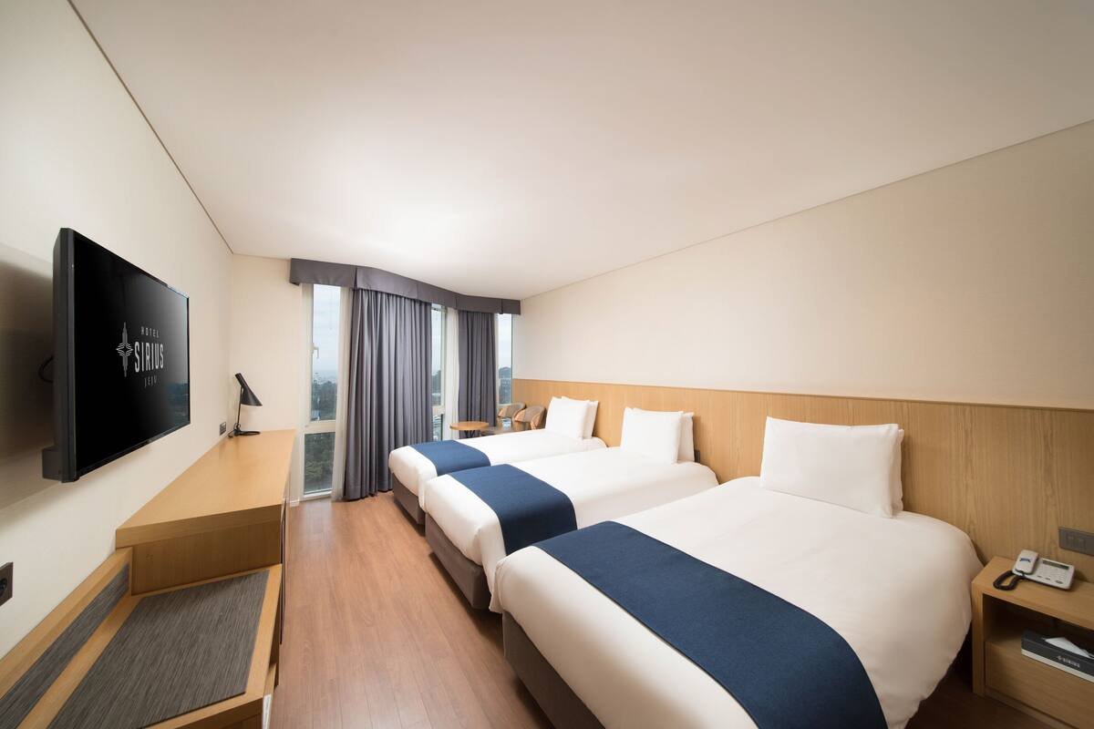 Habitación de hotel con tres camas, televisión, escritorio y ventana con cortinas.
