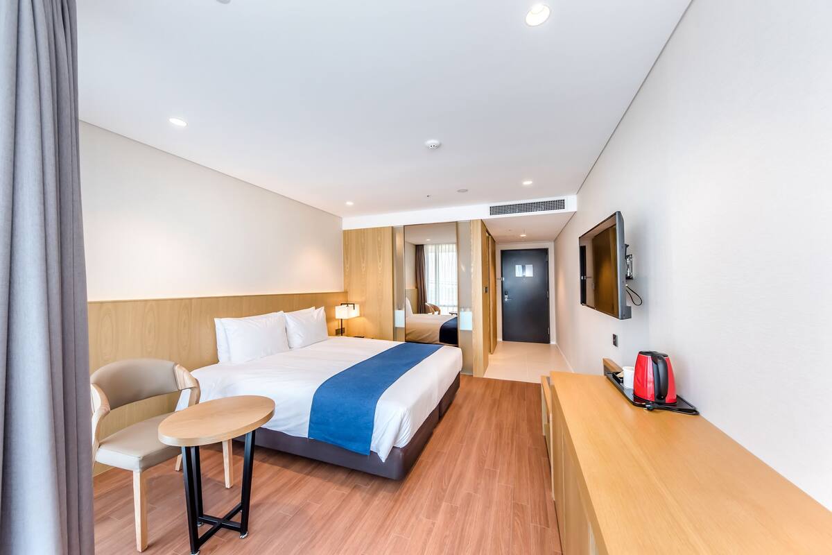 Una habitación de hotel moderna con cama, mesita de noche, silla y escritorio con tetera roja.