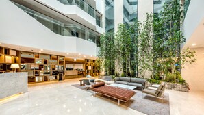 Un vestíbulo amplio con diseño moderno, que incluye una zona de estar con sofás y una butaca, un mostrador de recepción y un gran jardín interior con plantas altas.