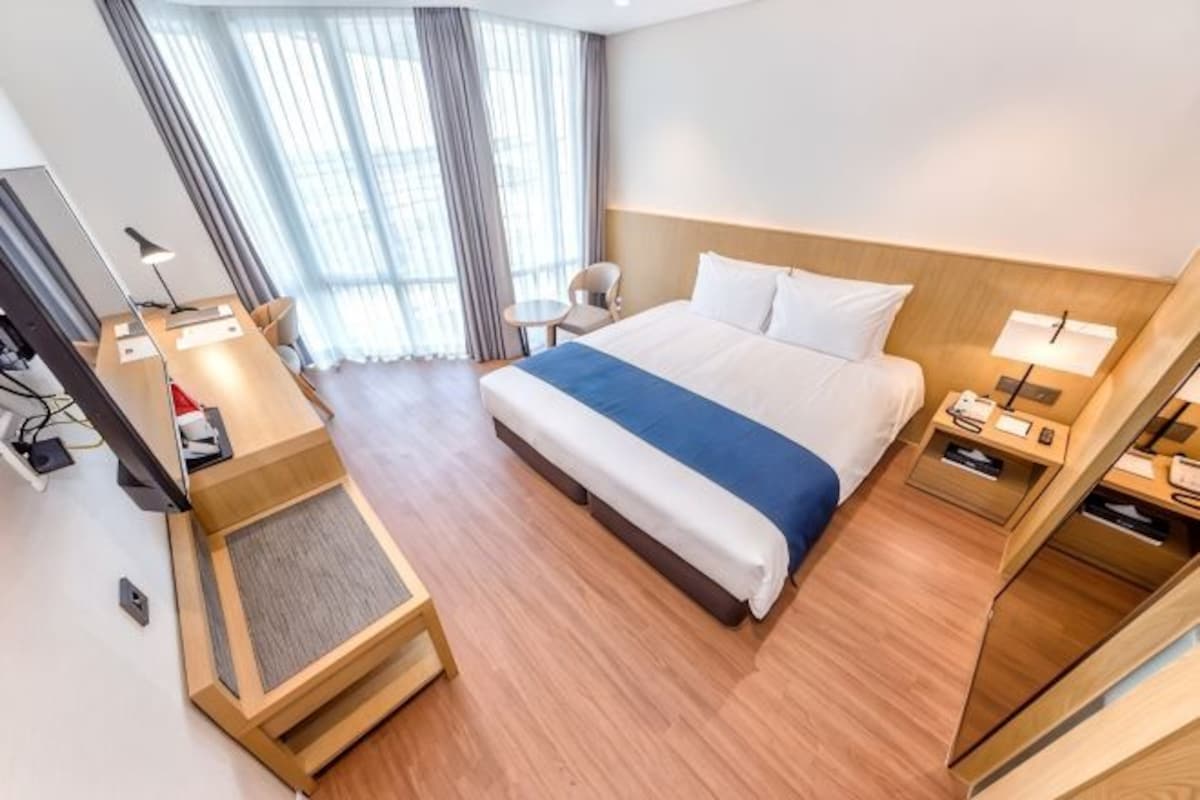 A hotel room with a bed, bedside tables, a desk with a chair, a small table, and a window with curtains.