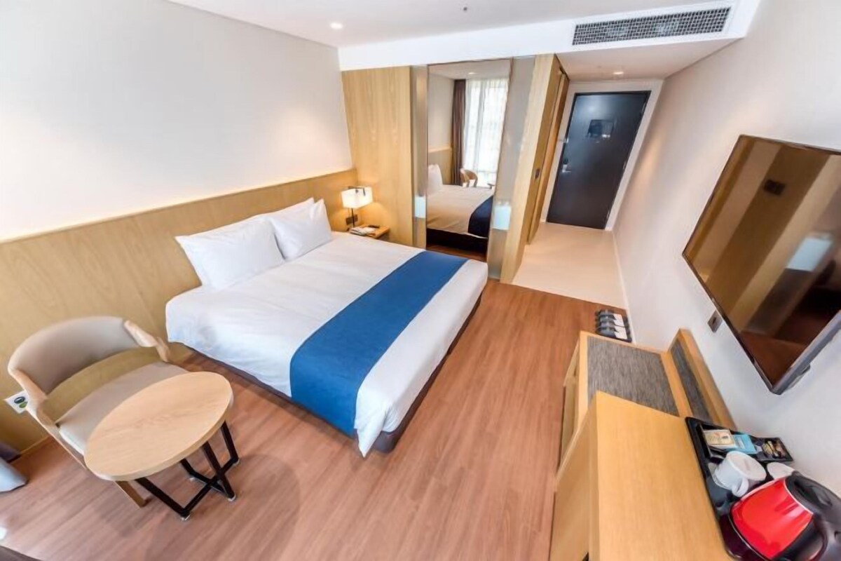Habitación de hotel con cama, una silla, una mesita, televisor y una puerta que da a otra habitación.