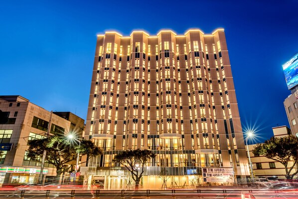 A modern high-rise building with a grid of windows, a well-lit facade, and a tree in front.