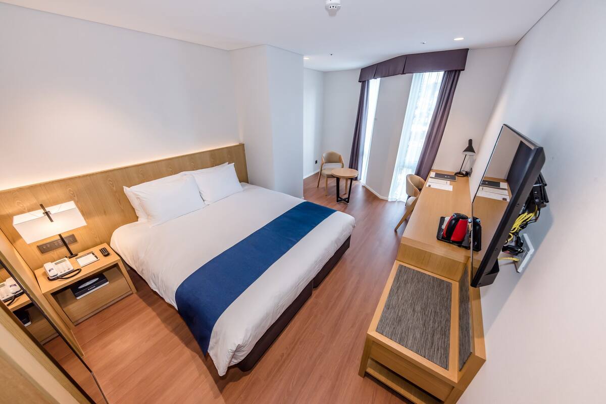 Habitación de hotel con una cama grande, un escritorio con computadora, una silla y una ventana con cortinas.
