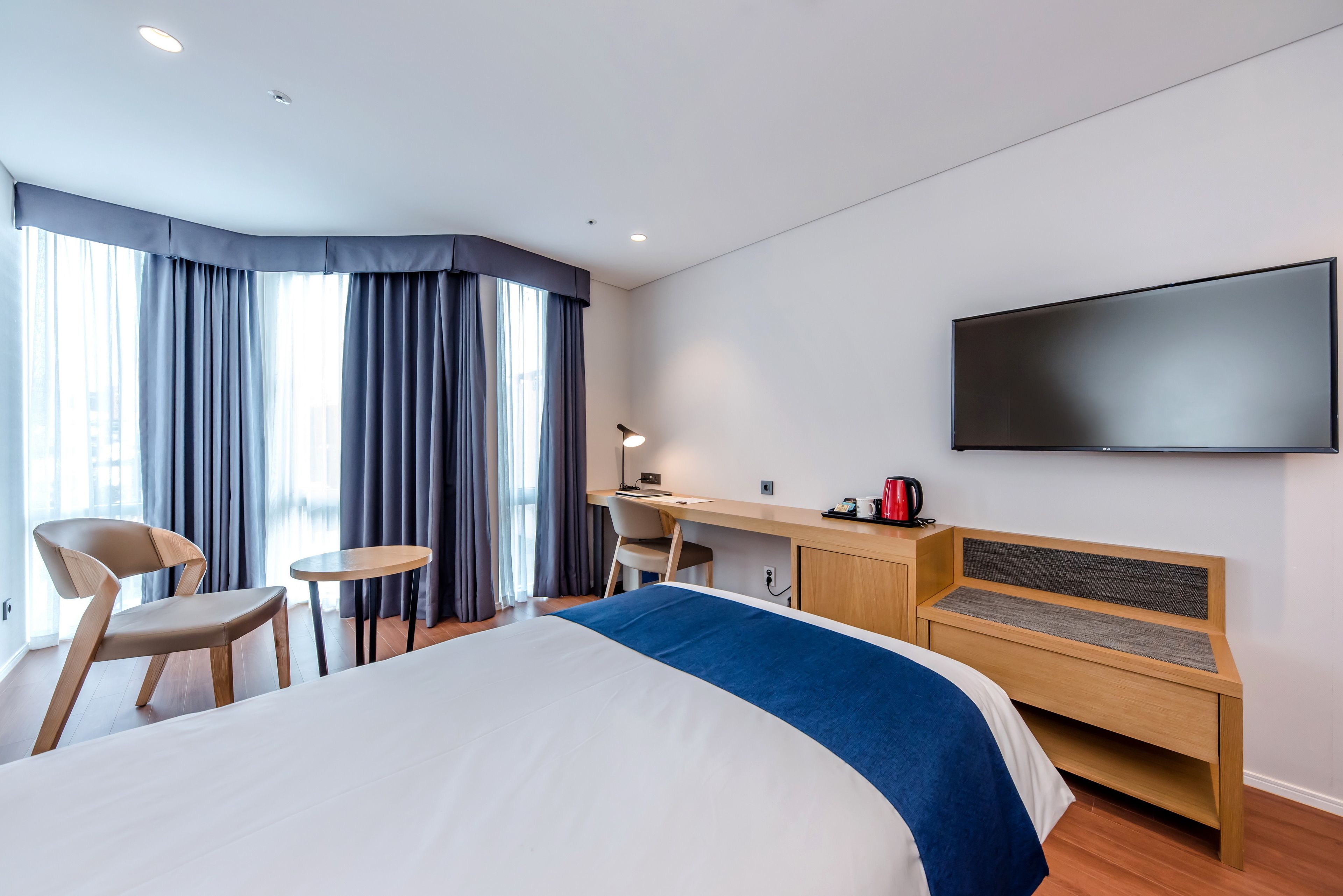 A modern hotel room with a large bed, a desk, a chair, a TV, and a view of the city through large windows.