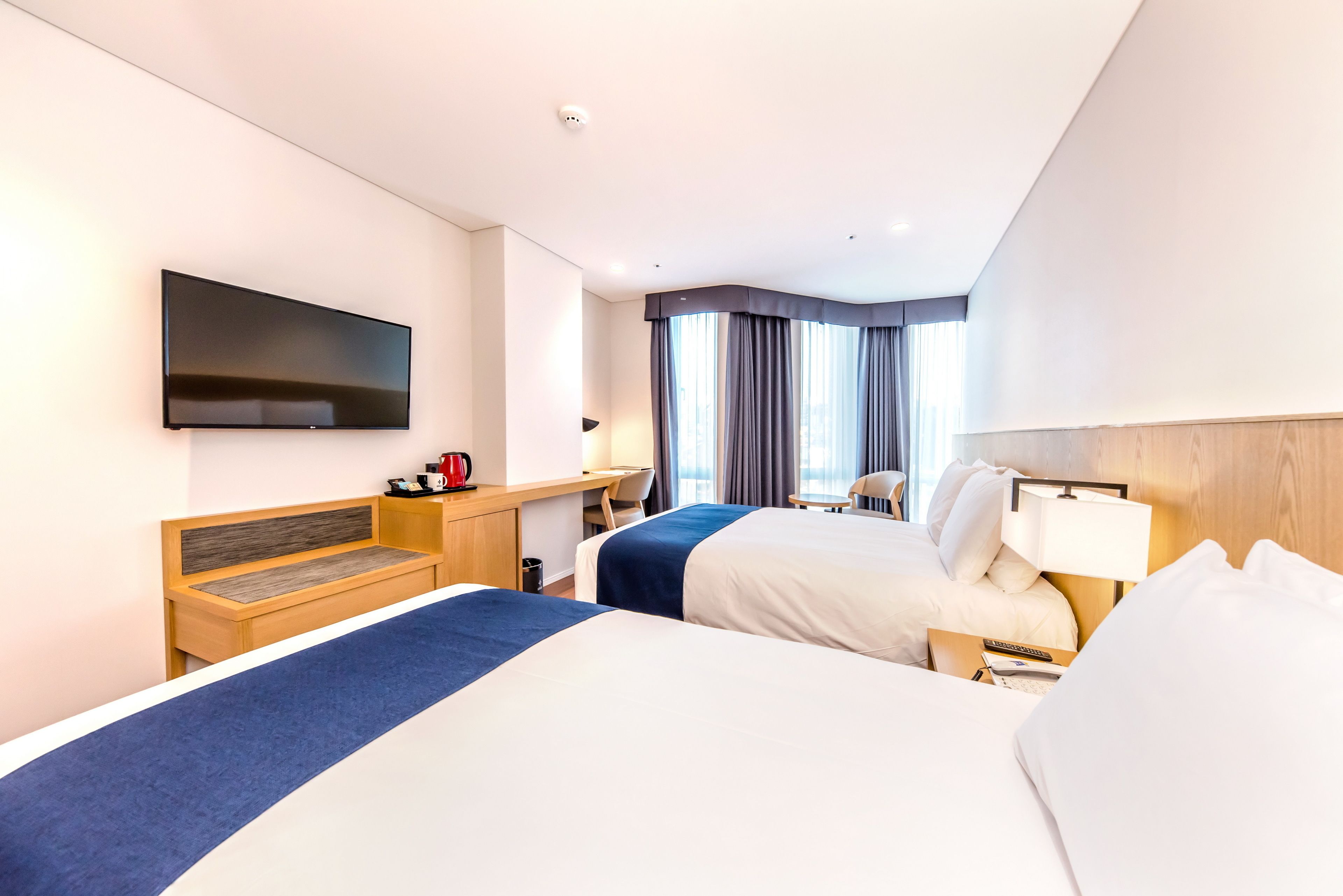 Habitación de hotel con dos camas, un televisor de pantalla plana montado en la pared, un escritorio con una silla, una ventana con cortinas y una mesita de noche con una lámpara.
