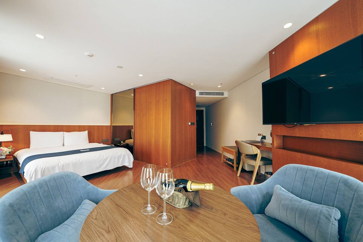 Habitación de hotel con cama, dos sillones, una mesa con vasos y una botella, y un televisor en la pared.