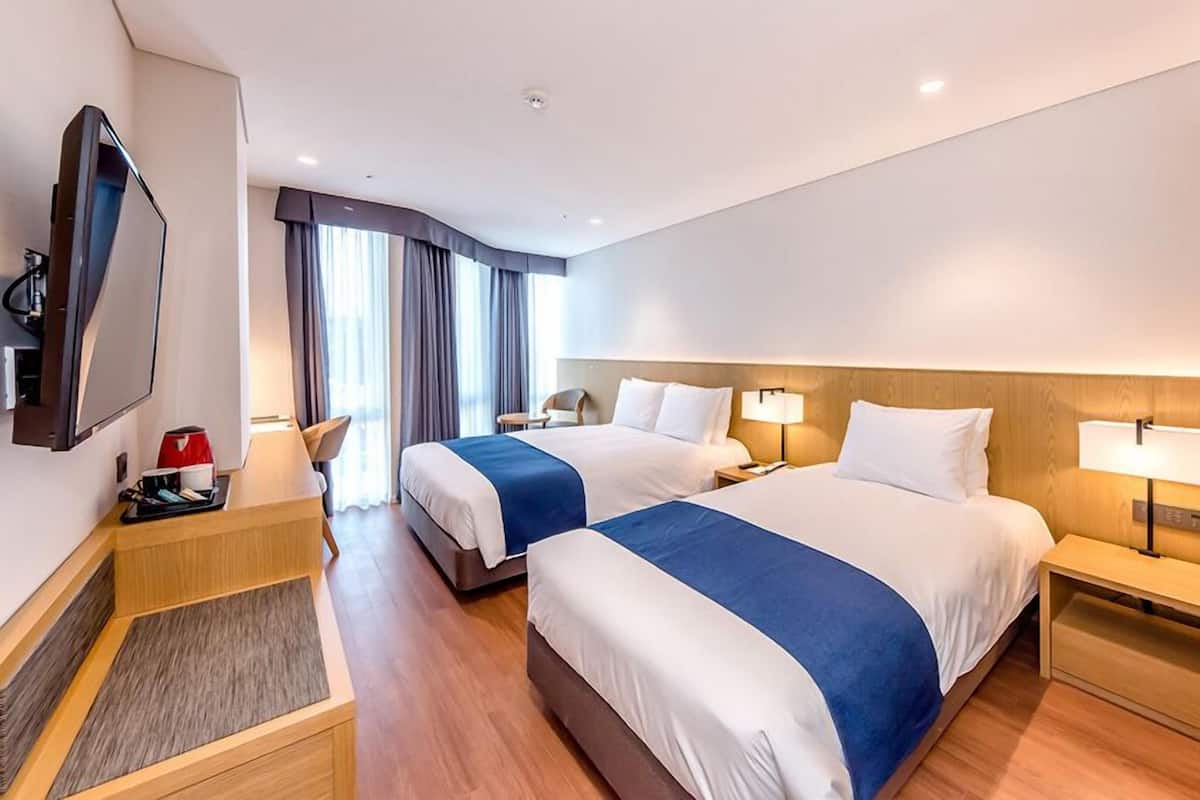Habitación de hotel con dos camas, un escritorio, televisor y ventana con cortinas.