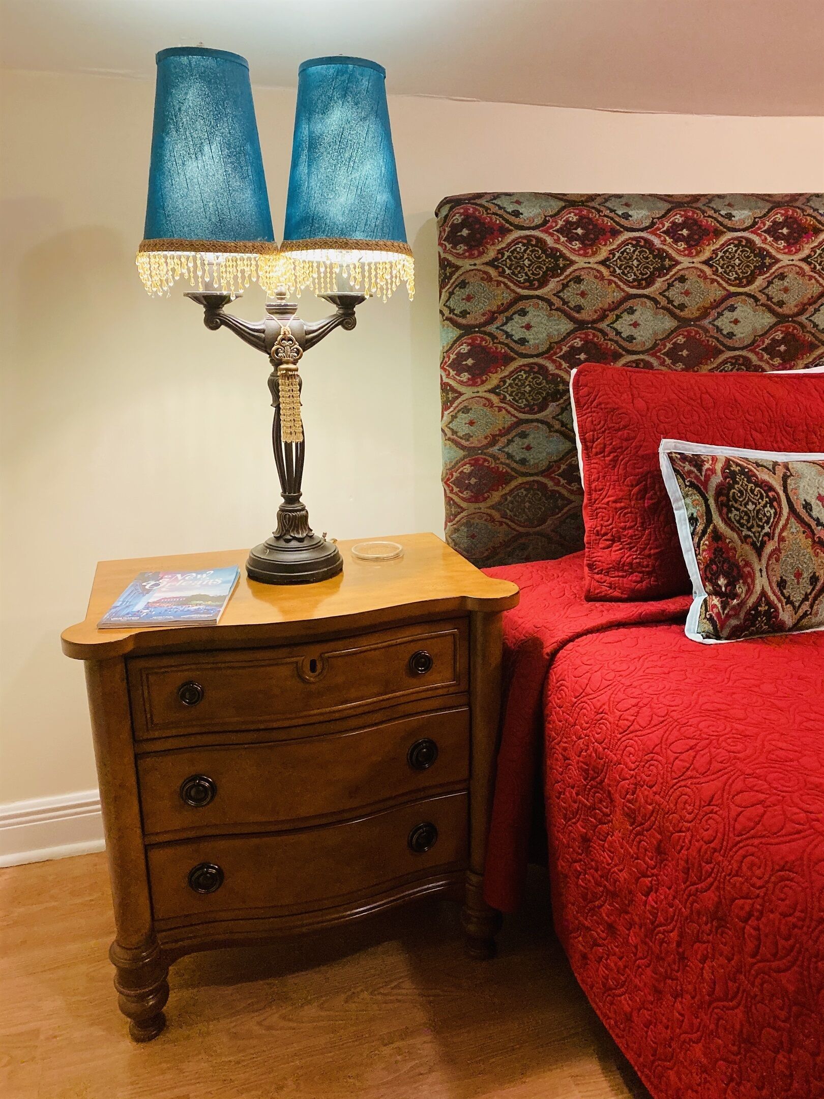 A wooden nightstand with a lamp, a red bed with patterned pillows, and a patterned headboard.