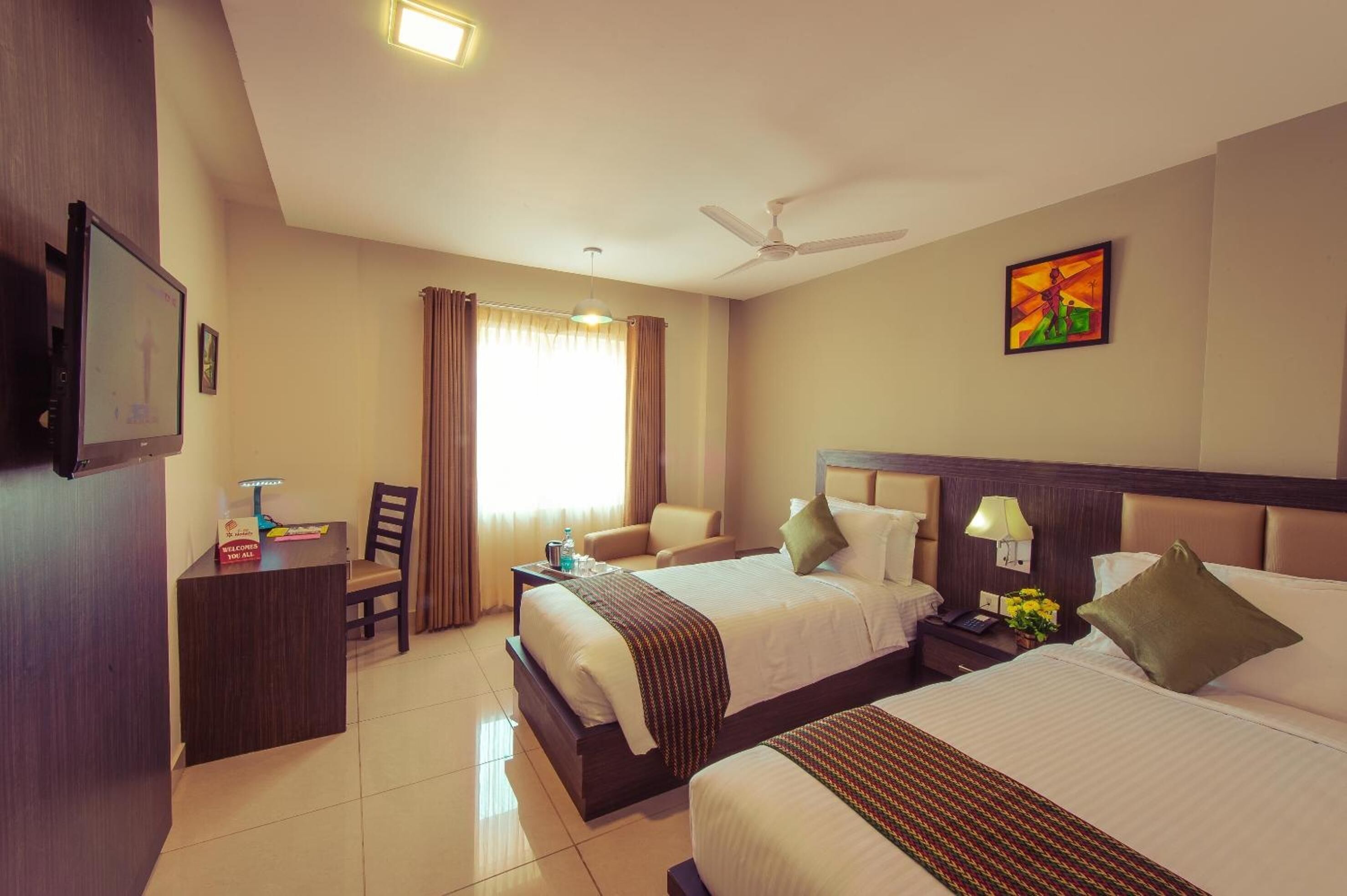 A hotel room with two beds, a desk with a computer, a chair, a TV, and a ceiling fan.