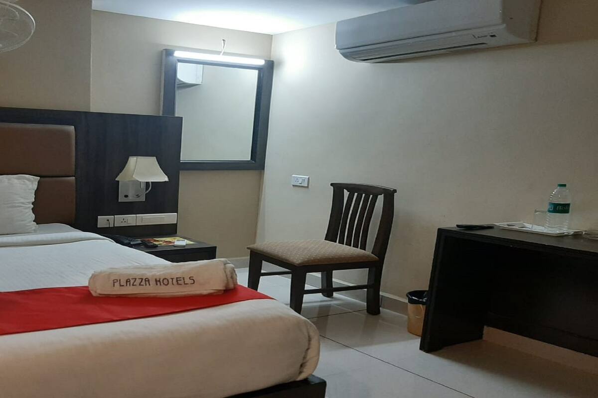 A hotel room with a bed, a chair, a desk, a lamp, and an air conditioner.