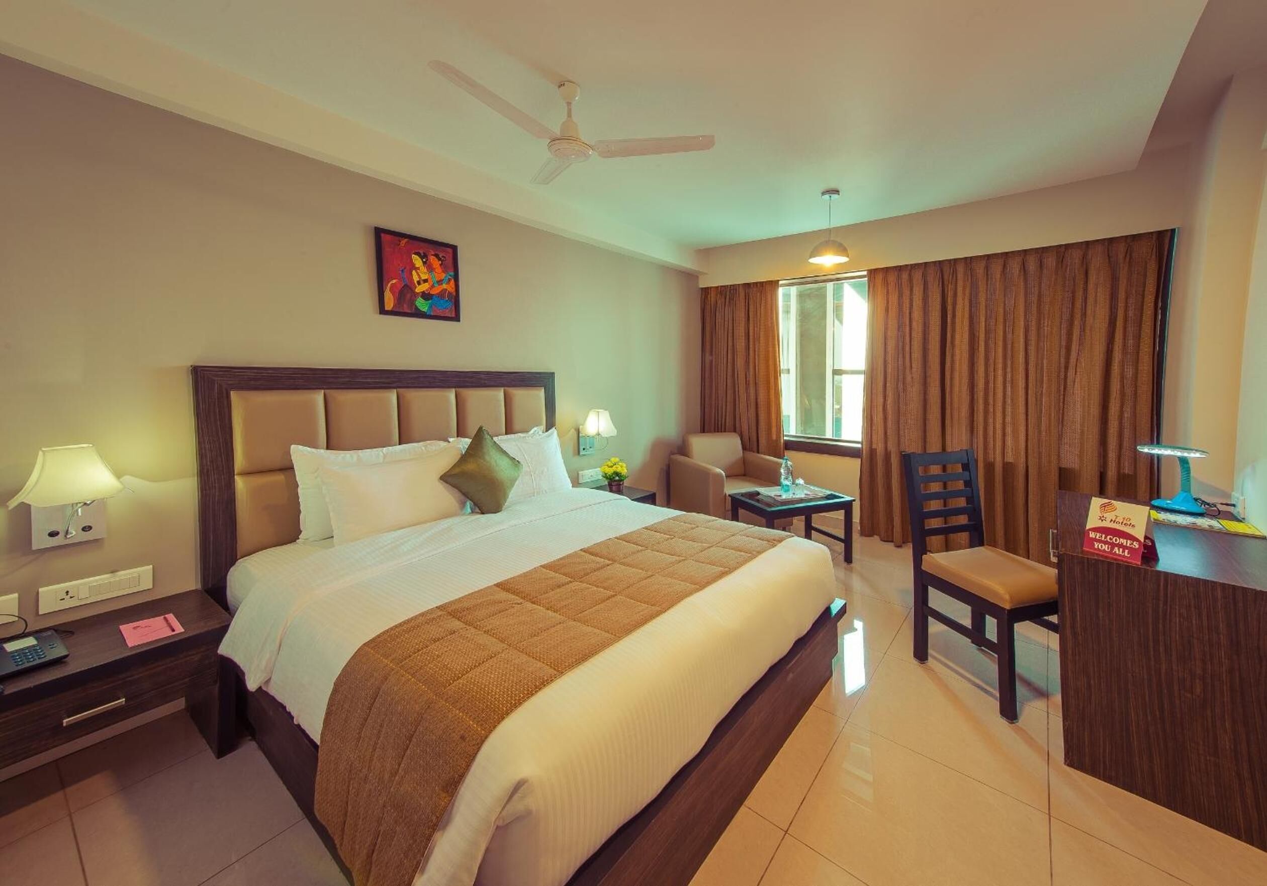 A hotel room with a large bed, a desk, a chair, a desk lamp, a nightstand, a phone, and a window with curtains.