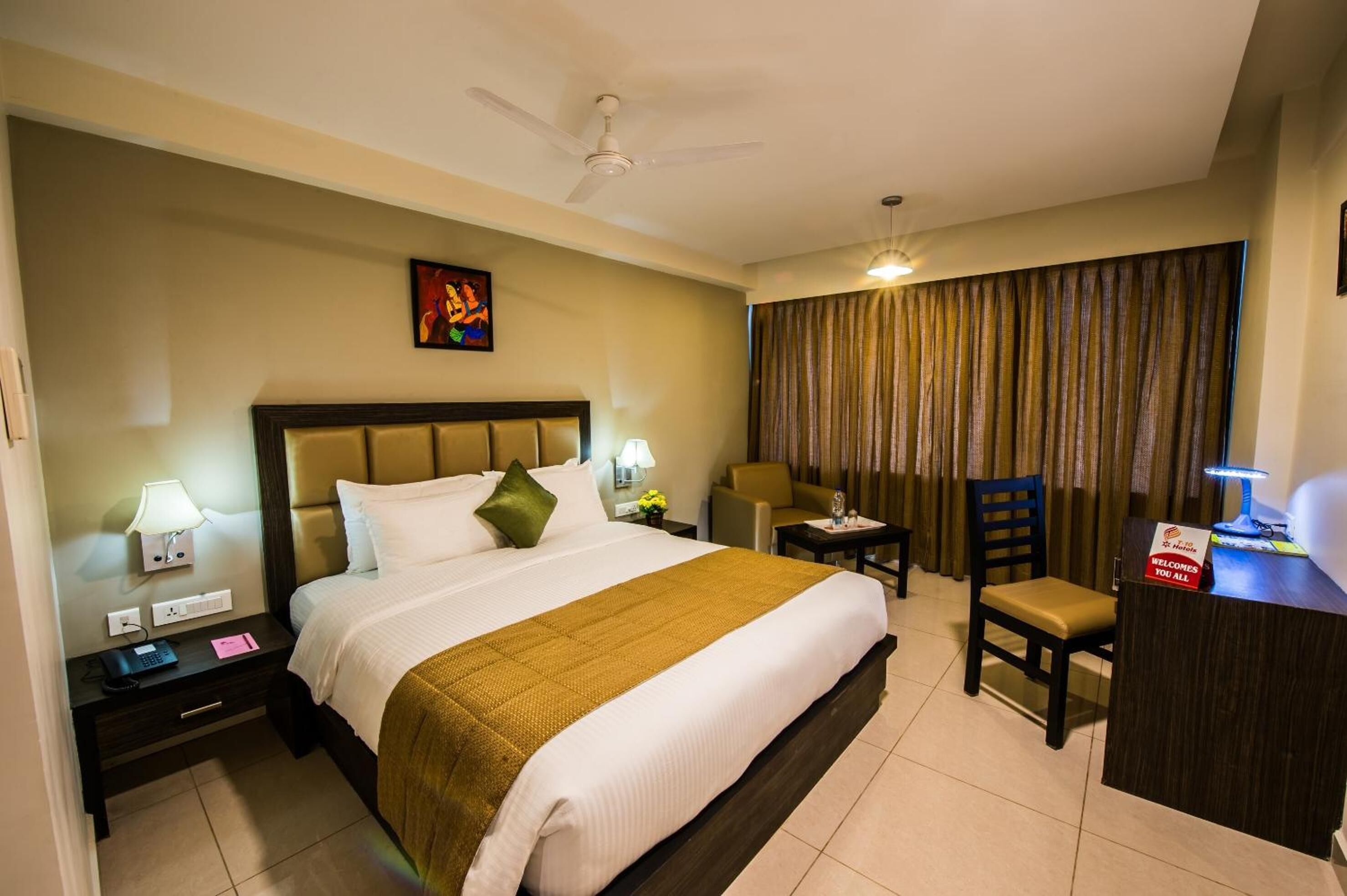 A hotel room with a bed, a desk, a chair, a nightstand, a lamp, a ceiling fan, and a curtain.