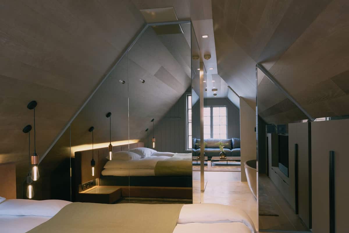 Ein modernes Schlafzimmer im Loft-Stil mit schrägen Wänden, einem großen Bett, einer Sitzecke mit einer Couch und Blick ins Freie.