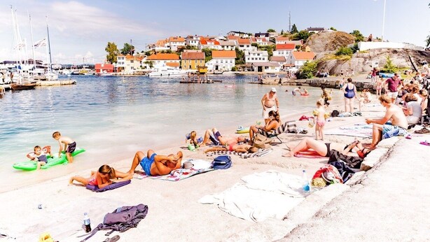 Une plage où des gens se prélassent au soleil et se baignent, un port de plaisance avec des bateaux, et des maisons en arrière-plan.