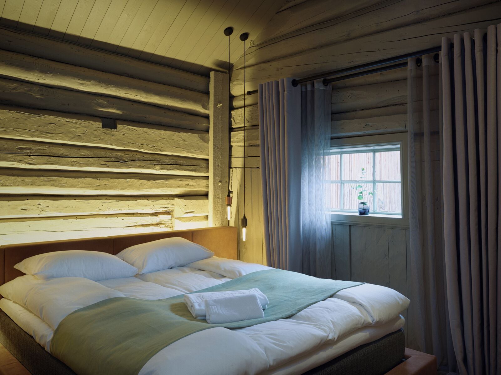 A wooden cabin-style room with a bed, curtains, and a window with a view.