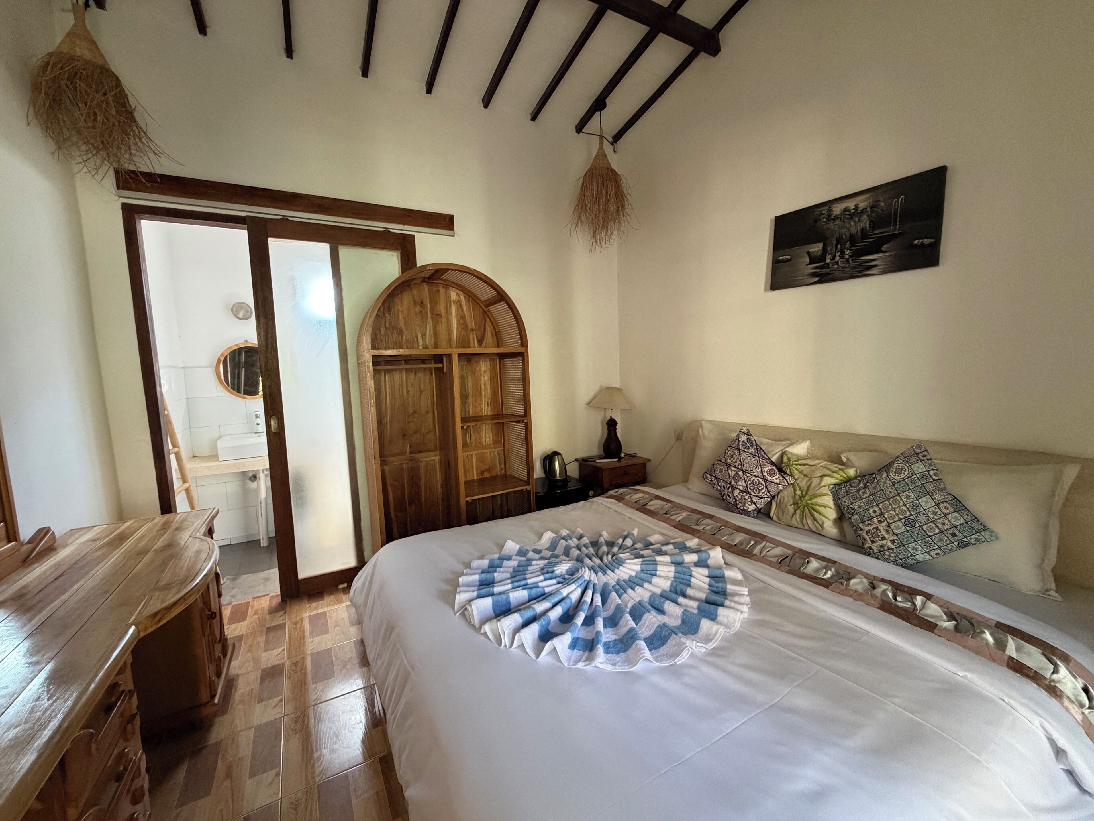A bedroom with a bed, a wooden wardrobe, a nightstand, a lamp, and a painting on the wall.