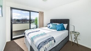 A bedroom with a large bed, a small side table, a view through a sliding glass door, and a ceiling light. - Accommodate Canberra - Century (Griffith)