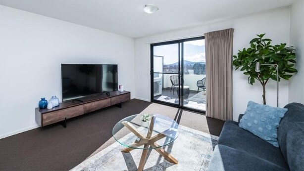A modern living room with a flat-screen TV, a glass coffee table, a sofa, a plant, and a balcony with a view of the city. - Accommodate Canberra - Century (Griffith)