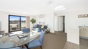 A modern living room with a glass dining table, blue chairs, a grey sofa, and a large window. - Accommodate Canberra - Century (Griffith)