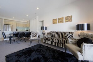 A modern living room with a black furry rug, a striped sofa, a dining area with a table and chairs, and a kitchen in the background. - Accommodate Canberra - Domain (Deakin)