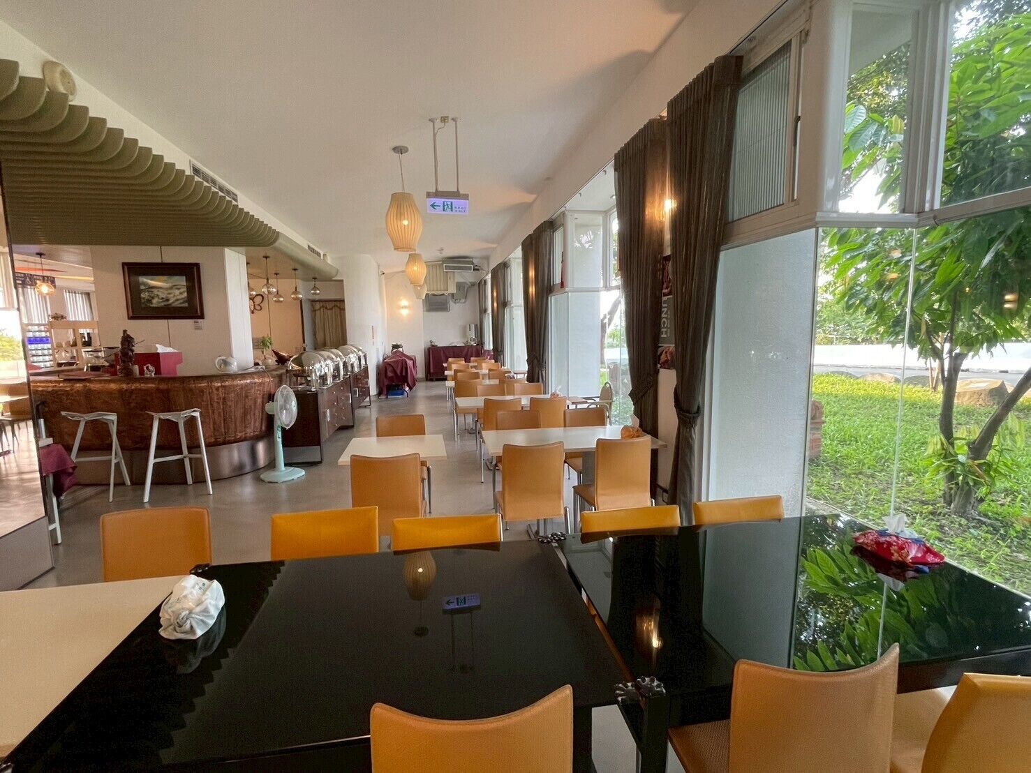 A spacious dining area with high tables and chairs, a bar area with stools, and large windows offering a view of greenery.